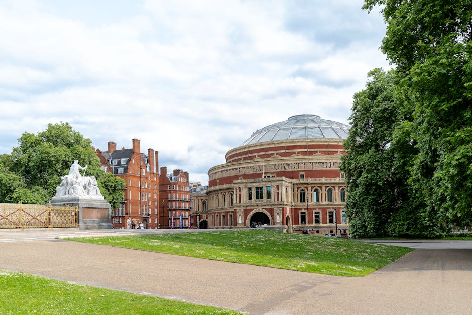 A wide view of a classical theatre building with a large, rounded brick facade and a domed roof, situated behind a grassy park area with paved walkways. To the left, there is a white marble statue of multiple figures on a stone pedestal, surrounded by a decorative metal fence. Tall, leafy trees flank the scene, providing greenery and shade. The sky above is partly cloudy, with some blue visible through the clouds, indicating daytime. The setting appears to be in an urban park or public space near historic buildings, aligning with the context of house removals and moving services offered by Man and Van Sydenham, who operate in the nearby area of Horniman Museum, Sydenham.