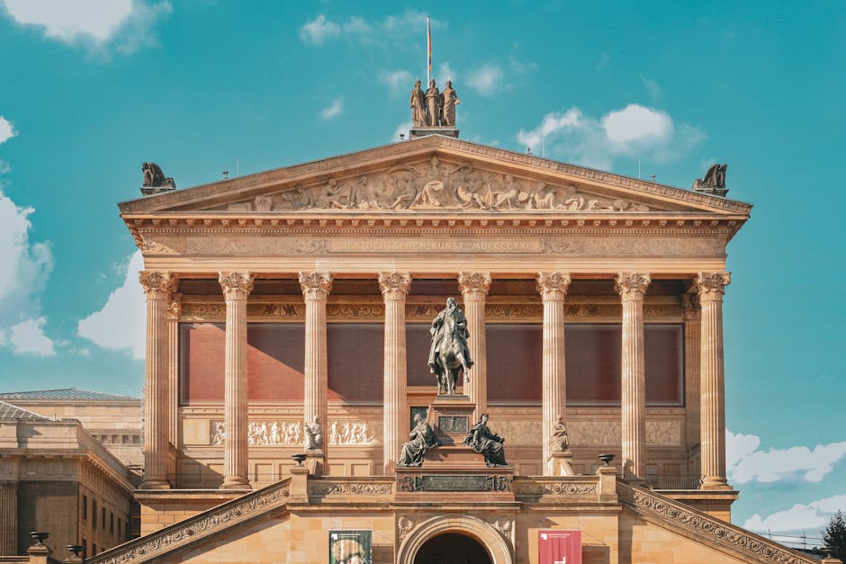 The image shows a classical-style building with a prominent triangular pediment supported by multiple tall Ionic columns. The pediment features detailed sculptural reliefs and is topped with a statue of four figures standing together. In front of the building, there is a bronze equestrian statue mounted on a pedestal, depicting a person on horseback. The building’s facade includes a balcony with decorative railings, and the materials used appear to be stone or marble with intricate carvings and inscriptions visible near the top of the building. The sky above is mostly clear with some scattered clouds, and the overall scene is well-lit, emphasizing the architectural details. This setting relates to a historic or cultural landmark, often associated with city centres or significant public spaces, relevant to the context of house relocations and moving services provided by Man and Van Sydenham, especially when transporting or displaying notable artifacts or collections.