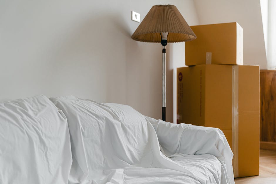 Inside a room prepared for home relocation, a white fabric-covered sofa is positioned against a light grey wall, with a tall brass floor lamp with a brown pleated lampshade standing nearby. To the right, three cardboard boxes of varying sizes, sealed with clear packing tape, are stacked in front of a wooden piece of furniture, possibly a cabinet or dresser, and are ready for moving. The boxes rest on a wooden floor, with natural light coming from a window to the right, partially visible with some sheer curtains. The scene suggests the packing and moving process, with furniture and boxes arranged for transportation, supported by the presence of a removal company's service, Man and Van Sydenham, involved in home relocation and furniture transport within the context of house removals near Sydenham, Lewisham.