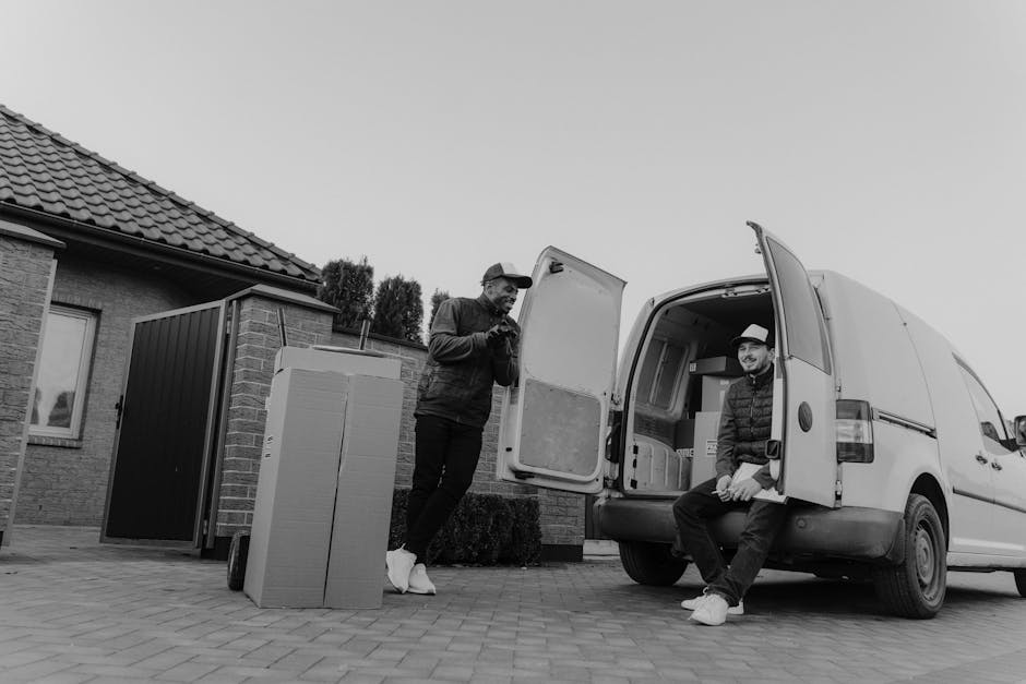 A man standing outdoors next to a white delivery van with its rear doors open, loading and stacking medium-sized cardboard boxes on the pavement during a home relocation process. The boxes are unseale