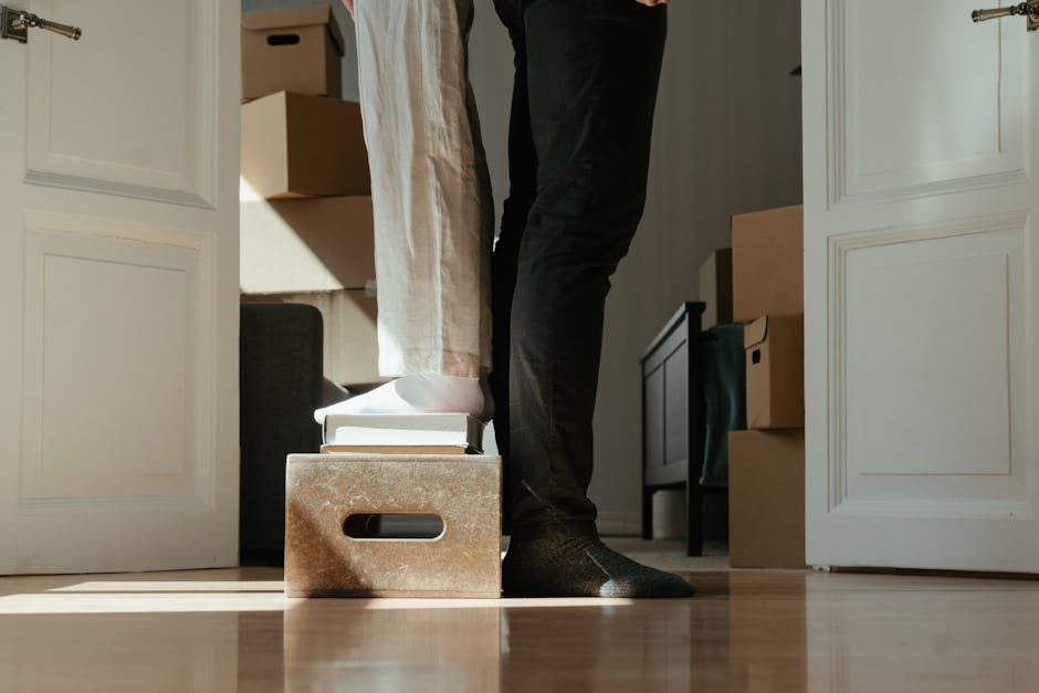 A person standing inside a room, lifting or positioning a cardboard box on top of a sturdy, brown, wooden storage box with a cutout handle, with a stack of books underneath. The scene appears to be part of a home relocation or packing process, as the individual is surrounded by moving boxes, some sealed with packing tape, and furniture such as a small dark table and a storage unit in the background. The room has white wooden doors or partitions on either side, and the floor is made of polished hardwood. Natural light illuminates the scene, highlighting the materials involved in packing and the careful handling of the items, which are likely being prepared for transportation by Man and Van Sydenham, specialists in removals and house moves, especially in areas with stairs like Sydenham flats.