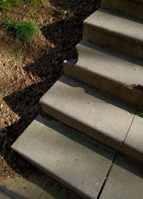 A close-up view of concrete stairs leading up to a residential property, with four visible steps bordered by a small patch of soil and some green grass on the left side. The stairs are outdoors, exposed to natural sunlight, casting shadows along the edges. The surface of each step is smooth, with slight variations in texture, and the edges are rounded. The surrounding area includes a dirt strip and a small area of greenery, indicating outdoor garden space. The image situates the stairs as part of a home entrance, relevant to house removals and moving services such as furniture transport or home relocation, with the focus on the steps that might represent a common point of access when moving large items or appliances into a property.