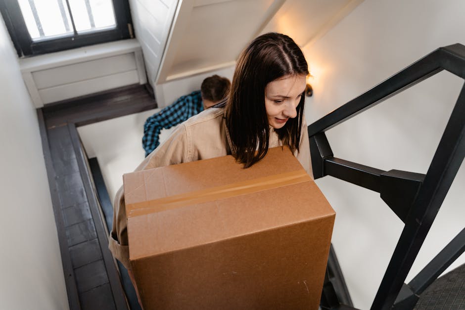 A person standing inside a room, lifting or positioning a cardboard box on top of a sturdy, brown, wooden storage box with a cutout handle, with a stack of books underneath. The scene appears to be part of a home relocation or packing process, as the individual is surrounded by moving boxes, some sealed with packing tape, and furniture such as a small dark table and a storage unit in the background. The room has white wooden doors or partitions on either side, and the floor is made of polished hardwood. Natural light illuminates the scene, highlighting the materials involved in packing and the careful handling of the items, which are likely being prepared for transportation by Man and Van Sydenham, specialists in removals and house moves, especially in areas with stairs like Sydenham flats.