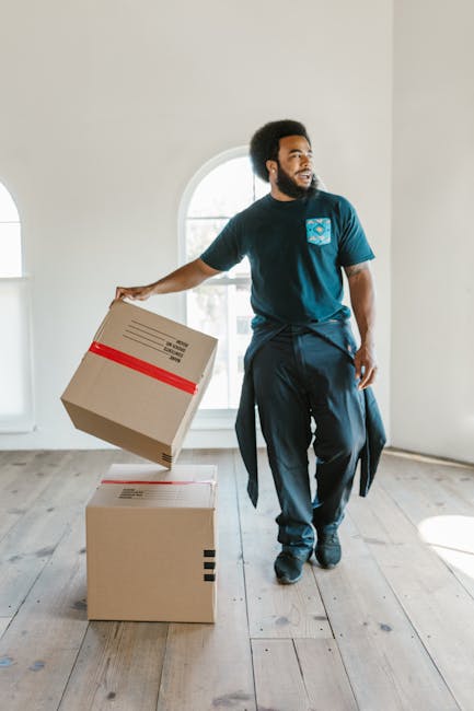 A professional mover from Man and Van Sydenham is inside a bright, spacious room with white walls and natural light streaming through a large arched window. He is wearing a navy-blue uniform with a company logo on the chest, and has a focused expression as he prepares for the home relocation process. The mover is holding a cardboard box sealed with red tape, lifting it over another box placed on the wooden floor. The room is empty aside from these boxes, indicating they are in the process of packing or unloading. The scene captures the systematic approach to furniture transport and packing involved in removals, with the mover demonstrating careful handling of the boxes, which are likely filled with household items to be relocated. This image illustrates the logistics and physical effort typical in professional moving services, and highlights the importance of organized packing during the loading process, with the manandvansydenham.co.uk team facilitating efficient furniture transport and home removals.