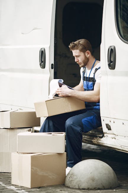 A man wearing a striped t-shirt and blue work overalls is seated on the edge of an open white van, surrounded by several cardboard boxes of varying sizes. He is holding a clipboard and appears to be checking or recording information related to the moving process. The boxes are stacked on the pavement beside him, with some placed inside the van, which is parked in an outdoor setting, possibly a driveway or street. The van’s sliding door is open, revealing part of the interior, and a small concrete bollard is visible nearby. This scene reflects the loading or packing phase of home relocation, typical of furniture transport and packing and moving activities, as might be managed by a professional removals service like Man and Van Sydenham.