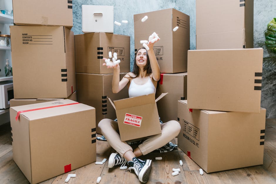 A professional mover from Man and Van Sydenham is inside a bright, spacious room with white walls and natural light streaming through a large arched window. He is wearing a navy-blue uniform with a company logo on the chest, and has a focused expression as he prepares for the home relocation process. The mover is holding a cardboard box sealed with red tape, lifting it over another box placed on the wooden floor. The room is empty aside from these boxes, indicating they are in the process of packing or unloading. The scene captures the systematic approach to furniture transport and packing involved in removals, with the mover demonstrating careful handling of the boxes, which are likely filled with household items to be relocated. This image illustrates the logistics and physical effort typical in professional moving services, and highlights the importance of organized packing during the loading process, with the manandvansydenham.co.uk team facilitating efficient furniture transport and home removals.