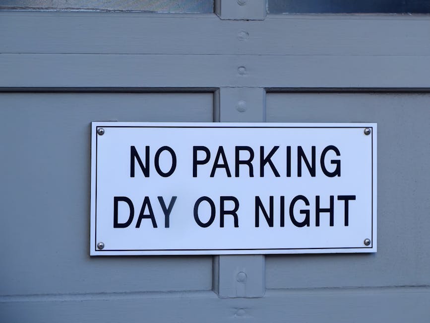 A rectangular white sign with bold black capital letters reading 'NO PARKING DAY OR NIGHT' is mounted on a grey surface, likely a garage door or wall, using four metal bolts at each corner. The surface appears to be painted wood or metal, with visible panel lines and a slight sheen, suggesting it is part of a building or property boundary. The lighting is natural and evenly distributed, illuminating the sign clearly. This signage is typically used to indicate parking restrictions in residential or commercial areas, and in the context of house removals or relocation services provided by Man and Van Sydenham, it highlights a parking regulation that may impact moving logistics and furniture transport near Sydenham. The presence of such signs is relevant for planning loading and unloading during home relocation or furniture transport to ensure compliance with local parking rules in Lewisham borough.