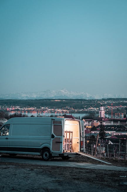 A grey panel van parked on an uneven, paved surface during dusk, with its side door open revealing a wooden furniture piece secured with straps inside, indicating a loading process for house removals. The van is positioned in an outdoor area with a view over a cityscape featuring multiple buildings, industrial structures, and distant mountains under a clear, twilight sky. The scene is lit naturally by remaining daylight, highlighting the materials used in packing and loading, such as cardboard boxes and protective blankets, which may be present nearby or inside the vehicle. This setup reflects the logistics involved in home relocation and furniture transport, with Man and Van Sydenham performing cargo handling and packing activities typical of a house moving service.