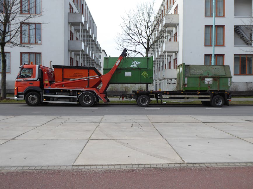 A flatbed truck operated by Man and Van Sydenham parked on a city street in front of residential apartment buildings during daytime. The truck has a red cab and a black chassis, with a hydraulic lifting arm extended to unload a large green metal skip container onto the street. Two additional large green skips, one with visible graffiti, are positioned on the flatbed for transport, ready for a house relocation or clearance. The street is paved with concrete slabs, and leafless trees line the sidewalk. The environment is overcast, providing diffused natural lighting suitable for documenting loading and transport activities involved in professional removals and moving services.