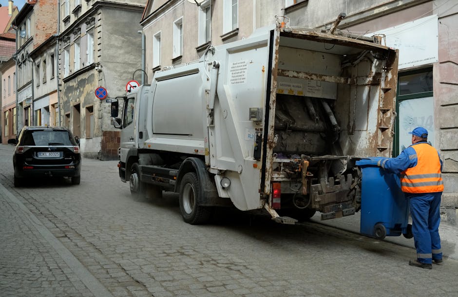 A sanitation worker wearing a blue uniform, orange high-visibility vest, and blue safety cap is seen emptying a blue wheeled bin into the rear opening of a large white garbage collection truck parked on a cobblestone street in an urban residential area. The truck belongs to Man and Van Sydenham, a professional removals and clearance service, and is positioned partially on the street and within a loading zone adjacent to a row of multi-story buildings with weathered facades. The open rear of the truck reveals rusted metal components and empty compartments designed for waste collection, with the truck's hydraulic lifting mechanism visible. Nearby, a parked black car is situated further down the street, and the scene is lit by natural daylight, depicting the logistics involved in house clearance, furniture transport, or home relocation activities, consistent with the services offered by [COMPANY_NAME].