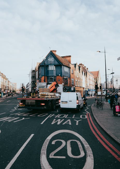 An image showing a busy street scene on Sydenham High Street SE26 with a flatbed truck parked in the middle of the road for furniture transport. The truck is loaded with various household items, including large cardboard boxes, wooden pallets, and wrapped furniture, positioned on its flat surface. A white van is parked alongside the truck, possibly assisting with the home relocation process. The background features a row of mixed architectural style buildings, some with brick facades and bay windows, lining the street. Pedestrians walk on the sidewalk, with bicycles parked nearby, and streetlamps line the pavement. The road has white and red markings indicating a 20mph speed limit and a mandatory directional lane, with an overcast sky overhead. The scene illustrates the logistics involved in furniture transport and loading during a house removal as handled by Man and Van Sydenham.