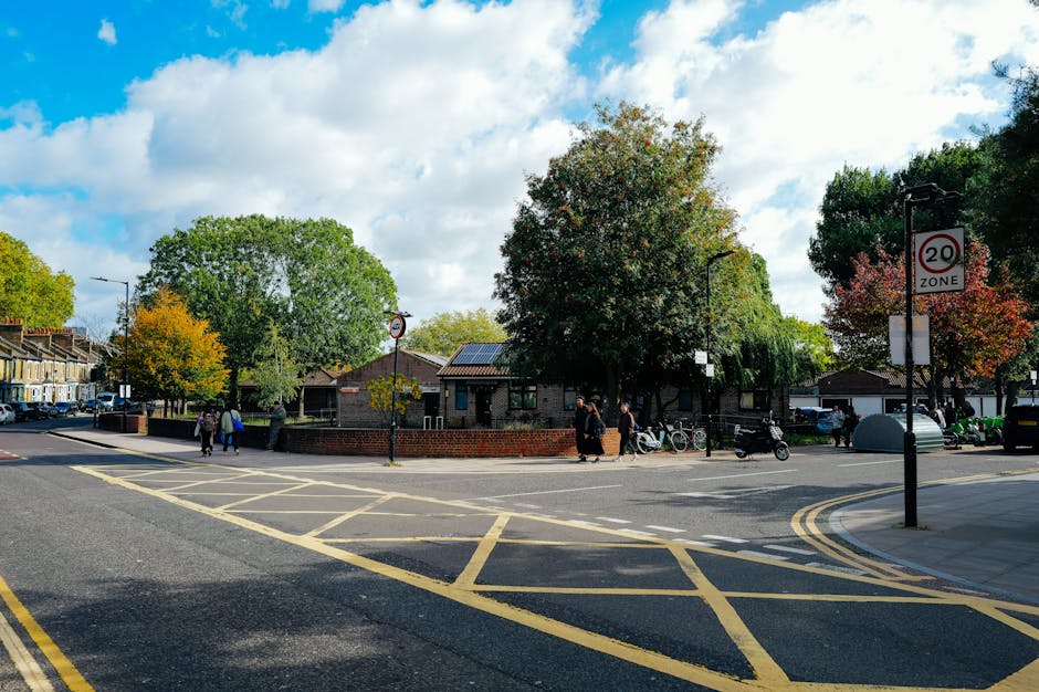 An image showing a busy street scene on Sydenham High Street SE26 with a flatbed truck parked in the middle of the road for furniture transport. The truck is loaded with various household items, including large cardboard boxes, wooden pallets, and wrapped furniture, positioned on its flat surface. A white van is parked alongside the truck, possibly assisting with the home relocation process. The background features a row of mixed architectural style buildings, some with brick facades and bay windows, lining the street. Pedestrians walk on the sidewalk, with bicycles parked nearby, and streetlamps line the pavement. The road has white and red markings indicating a 20mph speed limit and a mandatory directional lane, with an overcast sky overhead. The scene illustrates the logistics involved in furniture transport and loading during a house removal as handled by Man and Van Sydenham.