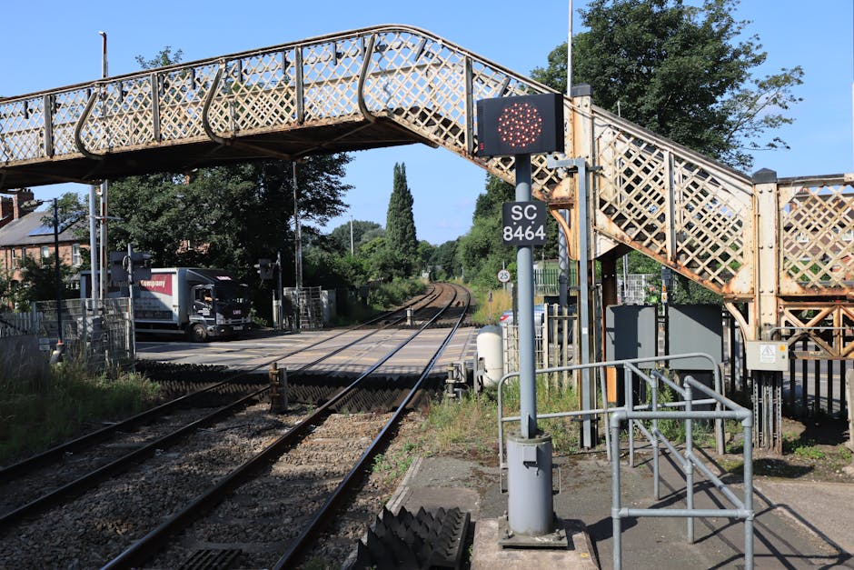 A London Underground train with red, blue, and silver exterior panels is stationary on the railway tracks at Sydenham station, visible from the platform. The platform is paved with concrete tiles and