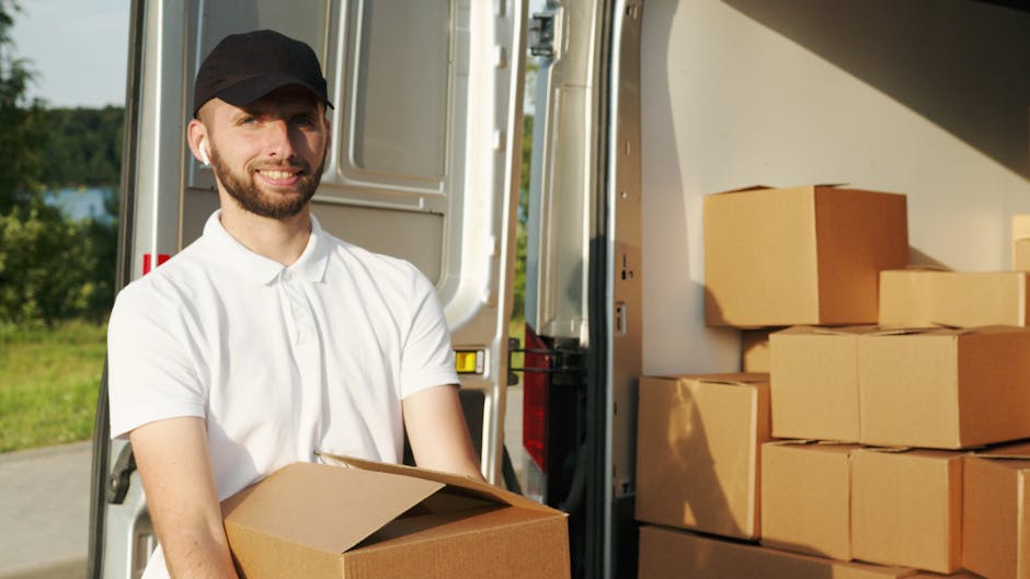 A man dressed in a white polo shirt and black cap is standing outdoors, smiling while holding a cardboard box. Behind him, there is a van with its back doors open, revealing an interior filled with neatly packed cardboard moving boxes of various sizes. Some boxes are stacked beside the van on the ground, ready for loading or unloading. The scene is set in a green outdoor area with trees and grass visible in the background, indicating a house removal or furniture transport process. The lighting suggests a sunny day, and the man appears to be involved in packing or unpacking during a home relocation, illustrating the logistical aspects of moving services offered by Man and Van Sydenham.