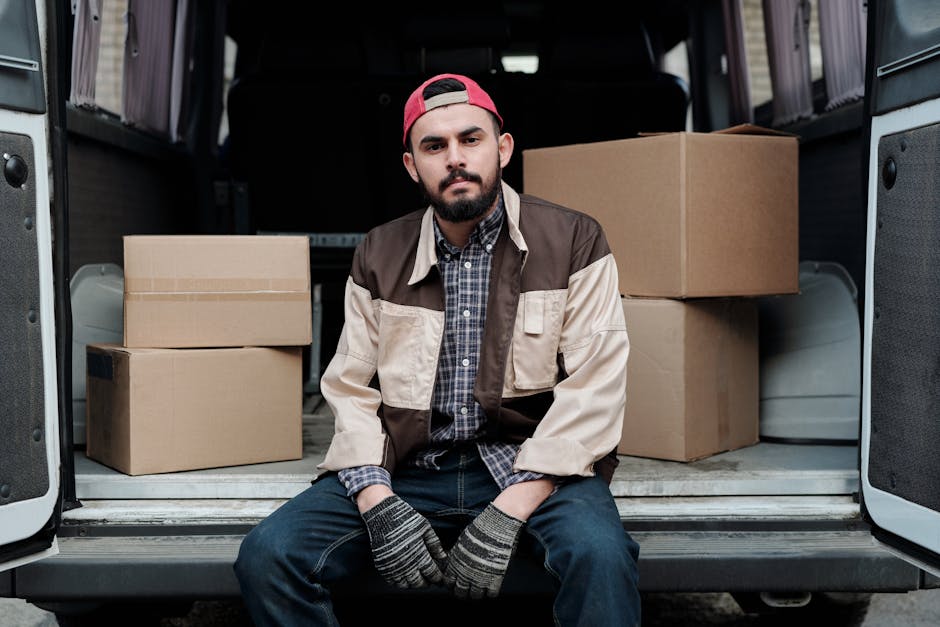 A man with a beard and mustache sitting on the edge of the open tailgate of a van during a home relocation, surrounded by several cardboard moving boxes of various sizes placed on the vehicle's floor inside. He is wearing a red cap backward, a beige and brown jacket over a blue plaid shirt, and striped gloves, indicating packing and handling activities. The background shows the dark interior of the van, with lighting illuminating the scene and highlighting the packing and transportation process involved in furniture transport or house removals, associated with services like those offered by Man and Van Sydenham, especially for last-minute moves in Sydenham.