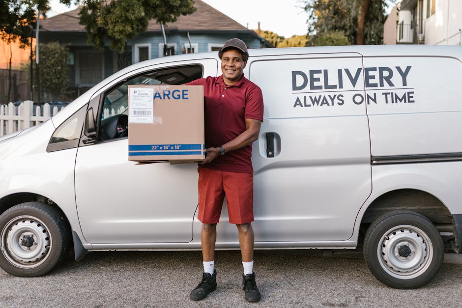 A man dressed in a white polo shirt and black cap is standing outdoors, smiling while holding a cardboard box. Behind him, there is a van with its back doors open, revealing an interior filled with neatly packed cardboard moving boxes of various sizes. Some boxes are stacked beside the van on the ground, ready for loading or unloading. The scene is set in a green outdoor area with trees and grass visible in the background, indicating a house removal or furniture transport process. The lighting suggests a sunny day, and the man appears to be involved in packing or unpacking during a home relocation, illustrating the logistical aspects of moving services offered by Man and Van Sydenham.